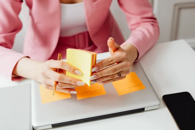 A woman in a pink blazer arranges sticky notes on her laptop, focusing on organization and planning. Checklista för dataskydd vid val av molntjänst