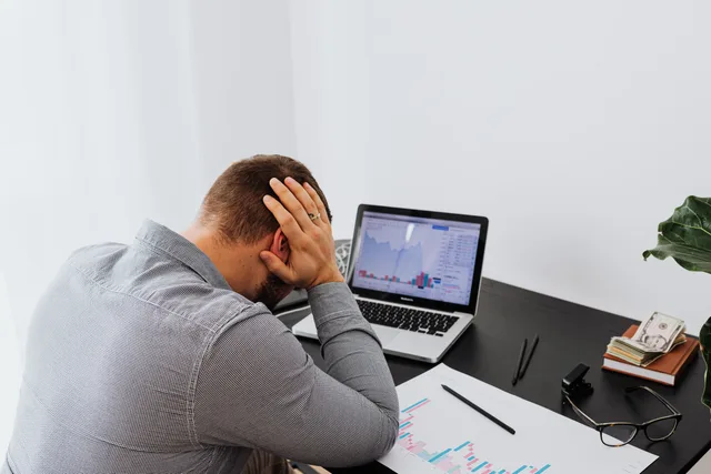 A businessman holds his head in frustration while sitting at a desk with a laptop and financial charts. Så utvärderar du programvara för svenska företag